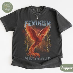 Può includere: T-shirt grigio scuro con un disegno di fenice. La parola "FEMINISM" è sopra la fenice, e "WE RISE FROM OUR ASHES" sotto. La fenice è rossa e arancione, con fulmini gialli.