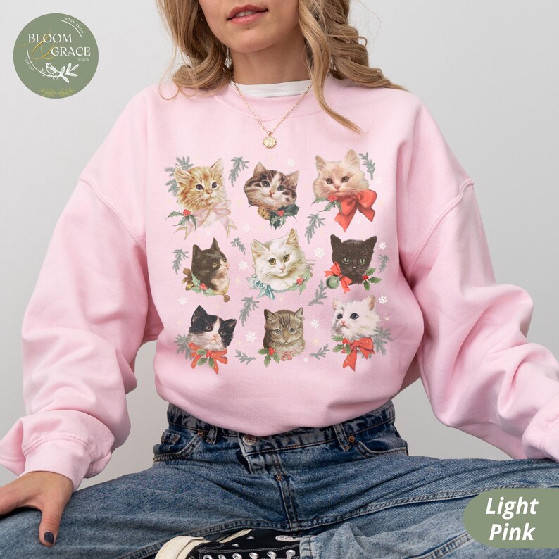 Peut inclure: Sweat-shirt rose clair orn&eacute; de portraits de chats avec des n&oelig;uds rouges et des d&eacute;corations festives. Le sweat-shirt a un col rond et des manches longues. Le logo Bloom & Grace est visible en haut &agrave; gauche. Les mots "Light Pink" sont en bas &agrave; droite.