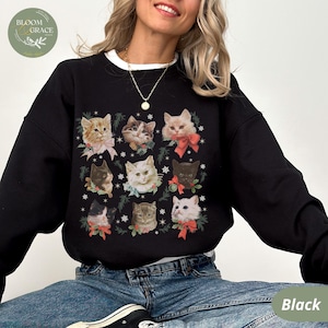 Peut inclure: Sweat-shirt noir avec un col blanc, orn&eacute; d'un motif de visages de chats avec des n&oelig;uds rouges et des brins de verdure. Le mot "Black" est visible en bas &agrave; droite.