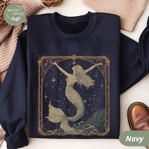 Puede incluir: Sudadera azul marino con una ilustración enmarcada en oro de una sirena con cabello suelto y cola escamada, sobre un cielo estrellado. La palabra "Navy" está impresa en la parte inferior. La sudadera es de color azul oscuro.