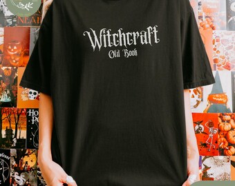 Op de afbeelding: Zwart T-shirt met de tekst "Witchcraft Old Book" in een gotisch lettertype. Het shirt is effen zwart en heeft een ronde hals. De afbeelding toont ook een gedeeltelijke weergave van een kleurrijke Halloween-achtergrond.