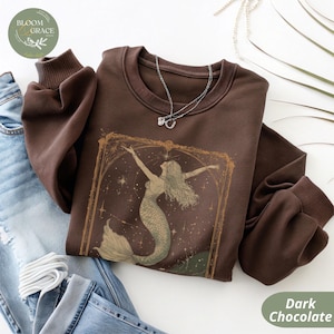 May include: Dark chocolate-colored sweatshirt featuring a mermaid graphic within a decorative frame. The design includes a mermaid with flowing hair and a fish tail, set against a starry background. The sweatshirt has a crew neck and long sleeves.