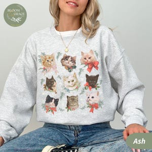Peut inclure: Sweat-shirt gris cendr&eacute; orn&eacute; d'un collage de neuf portraits de chats, chacun orn&eacute; de n&oelig;uds et de verdure festifs. Les chats varient en couleur et en motif, cr&eacute;ant un design charmant. Col rond et manches longues.