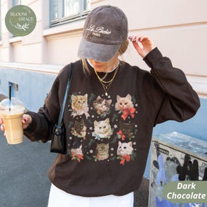 Peut inclure: Sweat-shirt chocolat fonc&eacute; avec une grille de neuf portraits de chats, chacun orn&eacute; de n&oelig;uds et de verdure festifs. Les chats varient en couleur et en motif. Le sweat-shirt est associ&eacute; &agrave; une casquette de baseball marron avec du texte blanc.