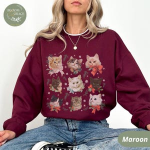 Peut inclure: Sweat-shirt bordeaux avec une grille de neuf portraits de chats, chacun orn&eacute; d'un n&oelig;ud ou de verdure festive. Les chats varient en couleur et en motif, sur fond de flocons de neige. Le sweat-shirt a un col rond et des manches longues.