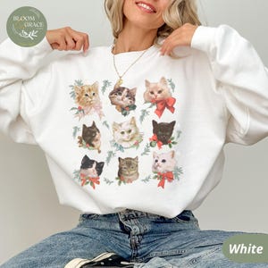 Peut inclure: Sweat-shirt blanc orn&eacute; d'une grille de neuf portraits de chats, chacun avec un n&oelig;ud ou une couronne. Les chats sont de diff&eacute;rentes couleurs, dont orange, noir et &eacute;caille de tortue. Le sweat-shirt a un col rond et des manches longues.