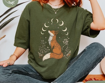 May include: A green t-shirt with a graphic of a fox looking up at a crescent moon with stars. The text "Bloom & Grace" is on the top left corner of the shirt. The shirt is made of hemp.