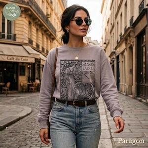 Puede incluir: Sudadera gris con un estampado gr&aacute;fico en blanco y negro y el texto "SPENSERS FAERIE QUEENE". Se combina con vaqueros azules y un cintur&oacute;n marr&oacute;n. La foto est&aacute; tomada en una calle adoquinada.