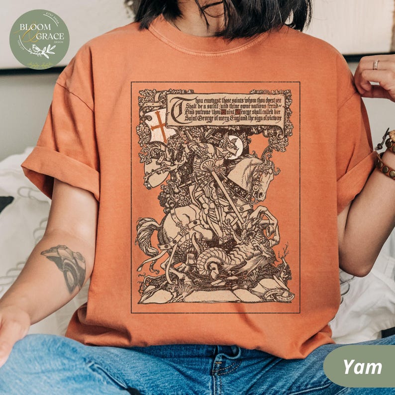 Pu&ograve; includere: T-shirt color igname con un'illustrazione seppia dettagliata di San Giorgio a cavallo che uccide un drago, con testo sopra. La maglietta ha maniche arrotolate e una vestibilit&agrave; rilassata.