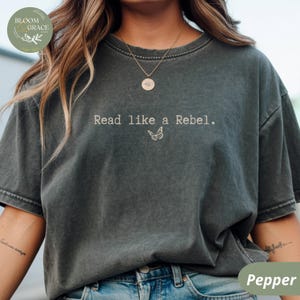 Könnte beinhalten: Dunkelgraues T-Shirt mit dem Text "Read like a Rebel" und einer Schmetterlingsgrafik. Das Shirt hat die Farbe Pfeffer und eine lockere Passform. Das Bloom & Grace-Logo befindet sich oben links.