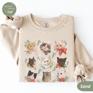 Peut inclure: Sweat-shirt couleur sable avec un motif de neuf chats, chacun orn&eacute; de n&oelig;uds et de verdure festifs. Les chats sont de diff&eacute;rentes couleurs et motifs, cr&eacute;ant un design charmant sur le th&egrave;me des f&ecirc;tes. Le sweat-shirt a un col rond et des manches longues.