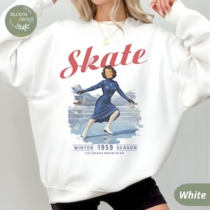 Puede incluir: Sudadera blanca con una ilustración vintage de una mujer patinando sobre hielo. La palabra "Skate" está en escritura roja encima de la imagen. Debajo de la imagen está el texto "Winter 1959 Season Colorado Mountains."