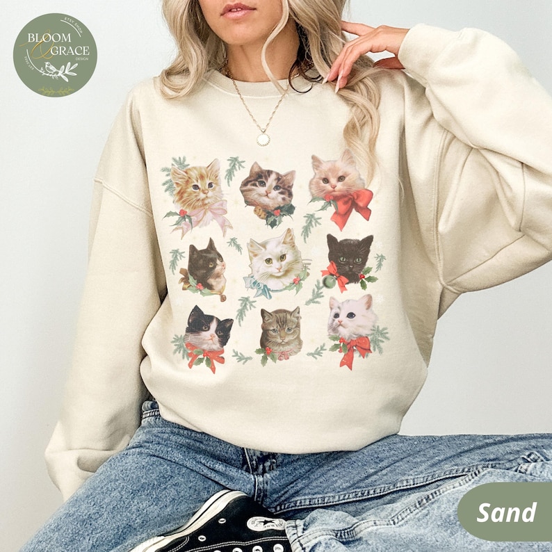 Peut inclure: Un sweat-shirt couleur sable orn&eacute; d'une grille de neuf portraits de chats, chacun orn&eacute; de n&oelig;uds et de verdure festifs. Les chats varient en couleur et en motif, cr&eacute;ant un design charmant sur le th&egrave;me des f&ecirc;tes. Le logo Bloom & Grace est visible en haut &agrave; gauche.