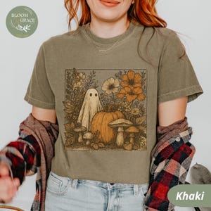 Pode incluir: Camiseta cáqui com uma ilustração em estilo vintage de um fantasma, abóbora, flores e cogumelos. O desenho é emoldurado dentro de um retângulo. A camisa tem mangas curtas e gola redonda. A palavra "Khaki" está impressa na parte inferior.