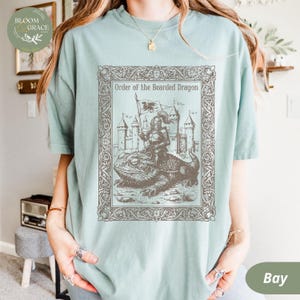 Puede incluir: Camiseta verde claro con una ilustración detallada de un caballero montando un dragón barbudo, con un castillo de fondo. El texto "Order of the Bearded Dragon" se muestra encima de la imagen dentro de un marco decorativo. La camiseta es de corte holgado.