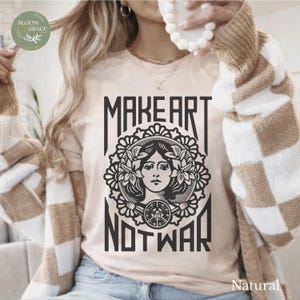 Op de afbeelding: Een beige t-shirt met een zwarte grafische print van een persoon omringd door bloemelementen en een cirkelvormig embleem. De tekst "MAKE ART" staat boven de afbeelding en "NOT WAR" staat eronder in een vetgedrukt, gestileerd lettertype.