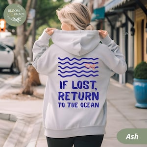 May include: Ash-colored hoodie with a drawstring hood. The back features a blue wave design with a peach-colored fish and the text "IF LOST, RETURN TO THE OCEAN" in blue. The hoodie is worn outdoors.