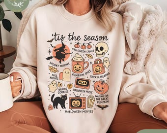 May include: Sand-colored sweatshirt with a Halloween-themed graphic. The design includes a witch on a broom, a skull, pumpkins, and the text "'tis the season." Other elements include ghosts, bats, and the words "Spooky Witches" and "Halloween Movies."