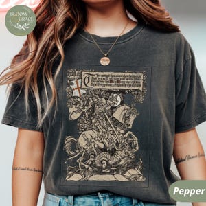 Pu&ograve; includere: T-shirt grigio scuro con un'illustrazione dettagliata di San Giorgio a cavallo che uccide un drago. Il design include testo in uno stendardo e una bandiera a croce. La maglietta ha un aspetto vintage.