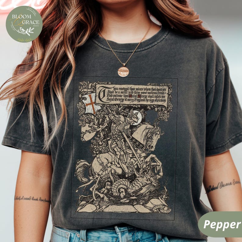 Pu&ograve; includere: T-shirt grigio scuro con un'illustrazione dettagliata di San Giorgio a cavallo che uccide un drago. Il design include testo in uno stendardo e una bandiera a croce. La maglietta ha un aspetto vintage.
