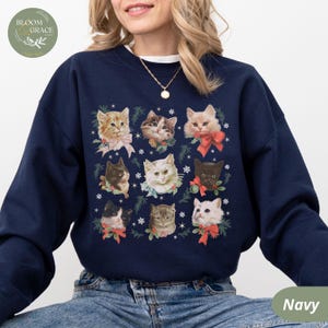 Peut inclure: Sweat-shirt bleu marine orn&eacute; d'un motif de neuf visages de chats, chacun orn&eacute; de n&oelig;uds, de verdure et de flocons de neige. Le sweat-shirt a un col rond et des manches longues. Le logo de la marque est en haut &agrave; gauche.