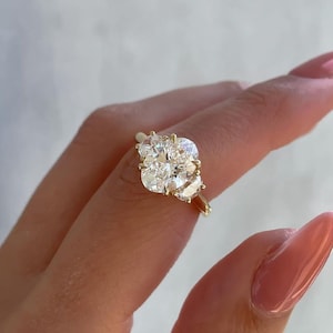 May include: A gold ring featuring a large, cushion-cut diamond center stone, flanked by two smaller pear-shaped diamonds. The ring is held up by a person's hand with pink nail polish.