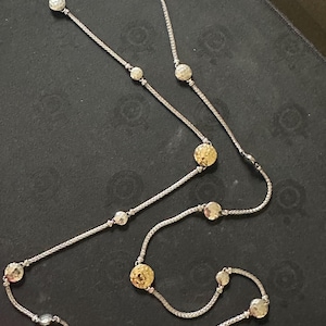 Pode incluir: Colar prateado com um design de corrente delicada. O colar é adornado com uma série de pingentes circulares texturizados em tons de prata e dourado. O colar tem um fecho de fecho.