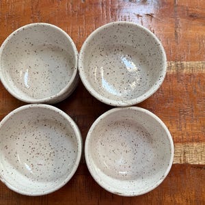 Op de afbeelding: Vier kleine, gespikkelde witte keramische kommen zijn gerangschikt op een houten oppervlak. De kommen hebben een eenvoudig, afgerond ontwerp en een gespikkeld patroon van donkerbruine stippen. De kommen zijn leeg en lijken nieuw.