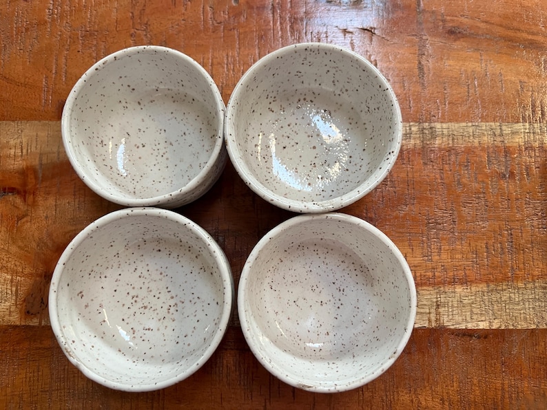 Op de afbeelding: Vier kleine, gespikkelde witte keramische kommen zijn gerangschikt op een houten oppervlak. De kommen hebben een eenvoudig, afgerond ontwerp en een gespikkeld patroon van donkerbruine stippen. De kommen zijn leeg en lijken nieuw.