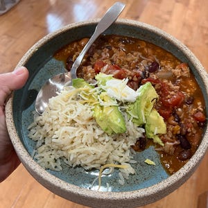 Può includere: Una ciotola di chili con riso, avocado, panna acida e formaggio grattugiato. Il chili contiene pomodori, fagioli e carne macinata. La ciotola è azzurra con un bordo strutturato e un cucchiaio d'argento all'interno.