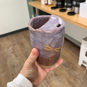 Può includere: Una tazza in ceramica fatta a mano con un design unico. La tazza presenta una combinazione di colori a due toni, con una parte superiore lavanda e una base marrone. Una libellula dorata è dipinta sul davanti. La tazza ha un ritaglio distintivo e un piccolo foro.