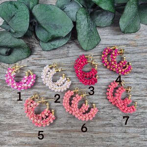 May include: A collection of beaded hoop earrings in various shades of pink, white, and red. Each earring features a gold-colored hoop and is adorned with small, colorful beads. The earrings are arranged on a wooden surface.