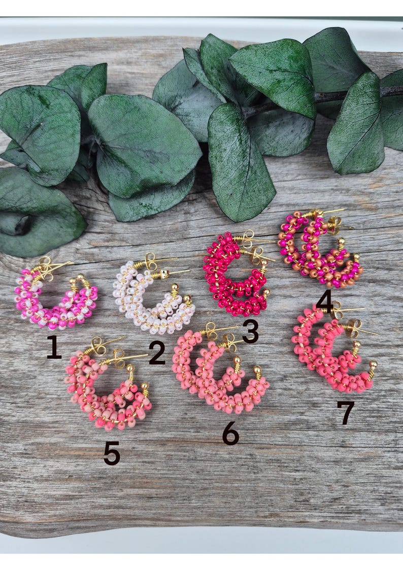 May include: A collection of beaded hoop earrings in various shades of pink, white, and red. Each earring features a gold-colored hoop and is adorned with small, colorful beads. The earrings are arranged on a wooden surface.