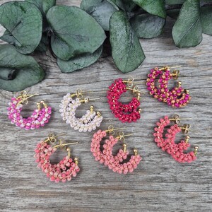May include: A collection of beaded hoop earrings in various shades of pink, white, and red, with gold-colored metal accents. The earrings are arranged on a wooden surface, with eucalyptus leaves in the background.