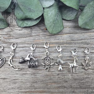 Könnte beinhalten: Ein Set aus sieben silberfarbenen Metallanhängern mit Karabinerverschlüssen. Die Anhänger haben verschiedene Formen, darunter ein Herz, ein gebogenes Horn, ein Blumenkübel, eine Kuckucksuhr, ein Pilz, ein stehendes Reh und eine sechsblättrige Blume.