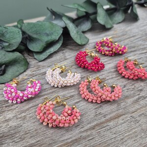 May include: A collection of beaded hoop earrings in various shades of pink and white. The earrings are arranged on a wooden surface, with gold-colored earring posts. Some earrings have a gradient effect, while others are solid colors.