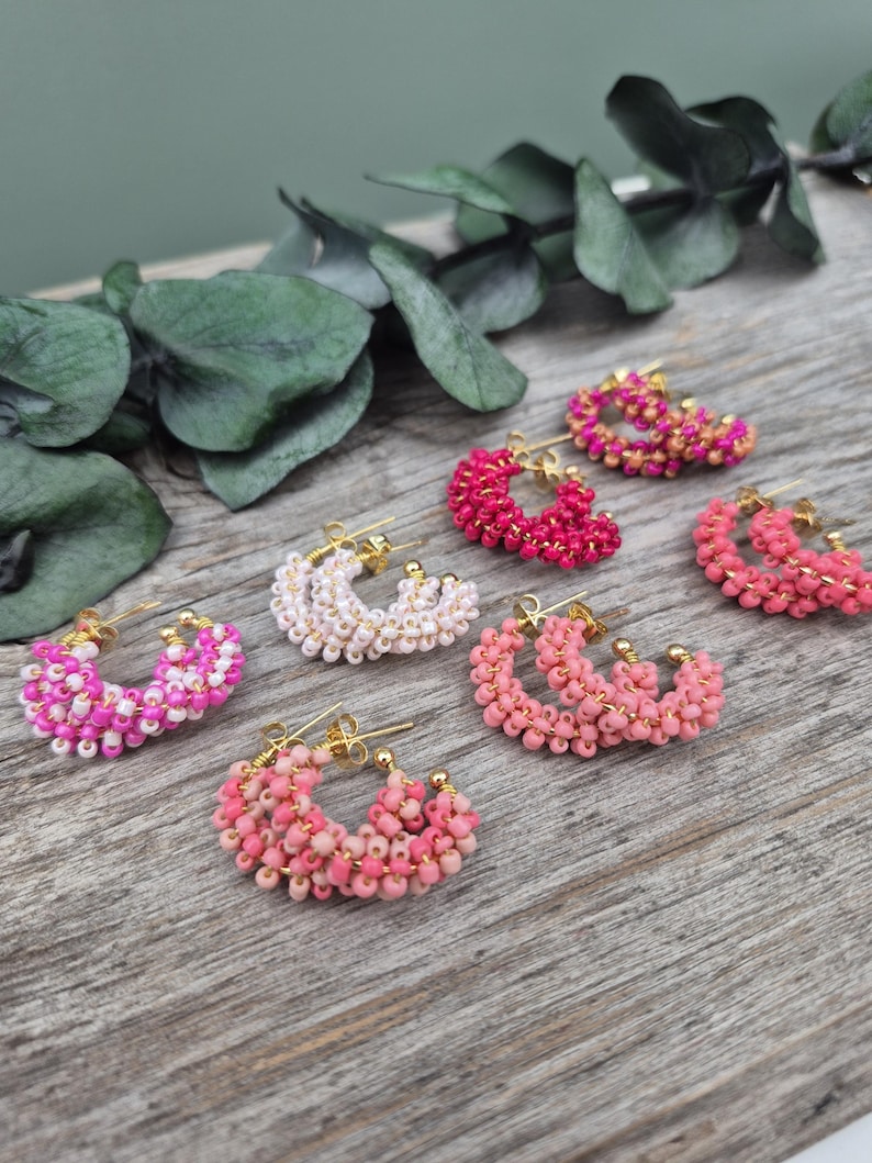May include: A collection of beaded hoop earrings in various shades of pink and white. The earrings are arranged on a wooden surface, with gold-colored earring posts. Some earrings have a gradient effect, while others are solid colors.