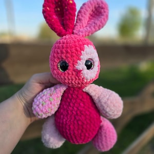May include: A handmade crochet bunny in shades of pink and white. The bunny has large, glittery eyes and long ears. The body is a solid magenta, with lighter pink and green accents on the paws and ears. A charming, soft toy.