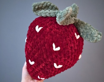 Peut inclure: Une grosse fraise en peluche rouge et verte. La fraise est faite d'une matière douce et texturée avec des détails de graines blanches. Les feuilles vertes sont attachées en haut.