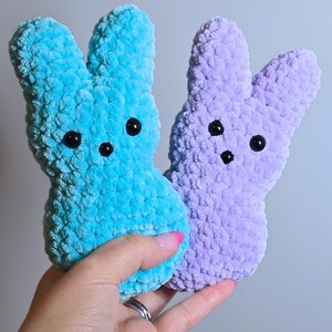 May include: Two crocheted bunny plushies, one in light blue and one in lavender. The bunnies have black eyes and a simple, smiling face.