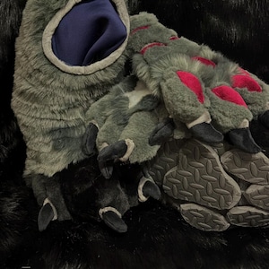 May include: A pair of gray and black furry monster paw gloves with red accents on the claws. The gloves have a dark blue interior and a textured sole. The gloves are designed to resemble a monster's paws.