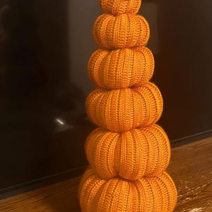 May include: A stack of six orange knitted pumpkins of varying sizes, arranged vertically. The pumpkins have ribbed detailing and are topped with a small twig and twine. The pumpkins are a fall-themed decoration.