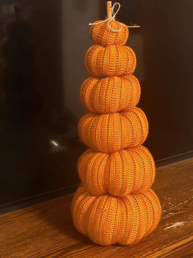 May include: A stack of six orange knitted pumpkins of varying sizes, arranged vertically. The pumpkins have ribbed detailing and are topped with a small twig and twine. The pumpkins are a fall-themed decoration.