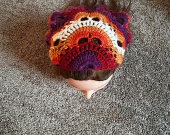 May include: A crocheted hat with a rainbow pattern in shades of purple, orange, red, and white. The hat has a scalloped edge and is made with a textured stitch.