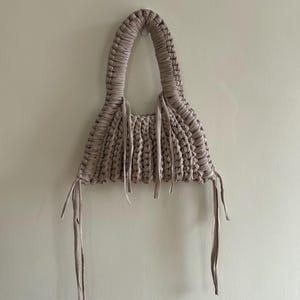 May include: A handmade, beige macrame handbag with a rounded handle and a triangular body. The bag features a woven design and long, dangling ties. The bag is hanging against a plain, off-white wall.