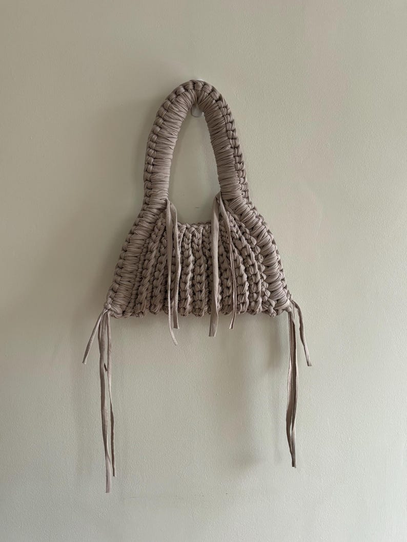 May include: A handmade, beige macrame handbag with a rounded handle and a triangular body. The bag features a woven design and long, dangling ties. The bag is hanging against a plain, off-white wall.
