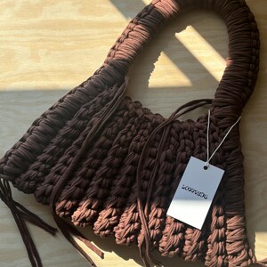 May include: A brown, handmade crochet bag with a curved handle. The bag features a textured, woven design and long, dangling ties. A white tag with the text "LOOPENDS" is attached to the bag.