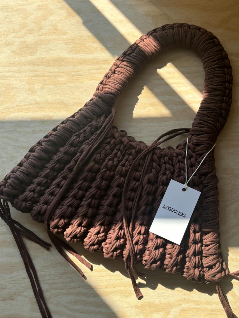 May include: A brown, handmade crochet bag with a curved handle. The bag features a textured, woven design and long, dangling ties. A white tag with the text "LOOPENDS" is attached to the bag.