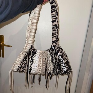 May include: Handmade macrame handbag in black, white, and beige. The bag features a woven design with a long handle and fringe detailing at the bottom. The bag is held by a person.