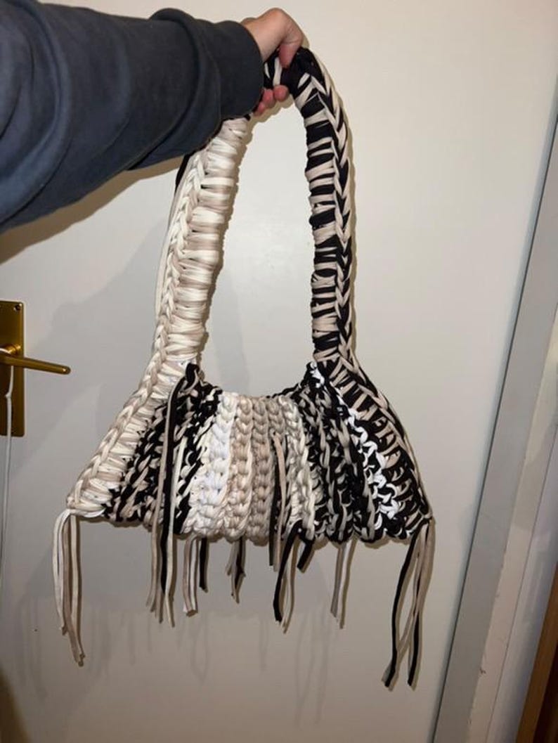 May include: Handmade macrame handbag in black, white, and beige. The bag features a woven design with a long handle and fringe detailing at the bottom. The bag is held by a person.
